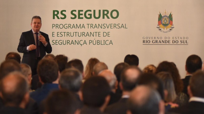 Fundo com imagem institucional do governo, pessoas de costas, vice-governador Ranolfo de pé com microfone na mão discursando.