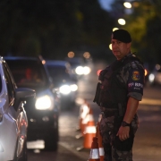 Em pé, policial militar observa o fluxo de veículos em barreira de trânsito montada pela 2ª Operação Integrada Metropolitana. À esquerda da imagem, fila de veículos.