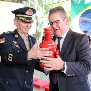 Governador em exercício segura miniatura de um hidrante que recebeu como homenagem na cerimônia de aniversário do batalhão. Ao fundo, à esquerda, soldados em fila. Ao fundo, à direita, bandeira do Brasil.