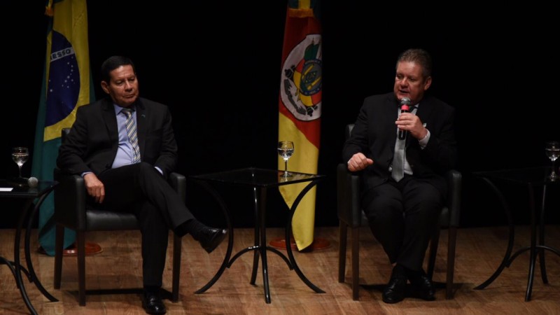 General Mourão e vice-governador Ranolfo sentados em cadeiras no palco do Teatro do Sesi. Ao fundo, bandeiras do Brasil e do RS.