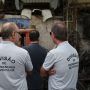 Dois servidores, de costas, com a inscrição "Divisão de Desmanches de Veículos" nas suas camisetas. Ao fundo, ferro-velho.