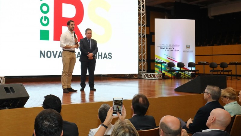 Em um palco, em pé, governador Eduardo Leite, com microfone na mão, ao lado do vice-governador Ranolfo Vieira Júnior. Ao fundo, telão com a logomarca do governo. RS.GOV Novas façanhas.