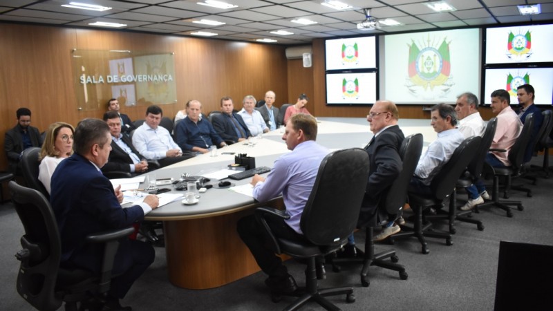 Ranolfo Vieira Júnior sentado à ponta de uma grande mesa de reuniões, rodeada por prefeitos da região do Alto Uruguai, na Sala de Governança do CAFF. Ao fundo, telões com o brasão do Estado.