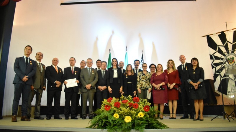 Os 15 homenageados com a Medalha Tiradentes e demais autoridades no palco do auditório do Palácio da Polícia, na Capital. À frente, arranjo de flores. À direita, banner com brasão da Polícia Civil.