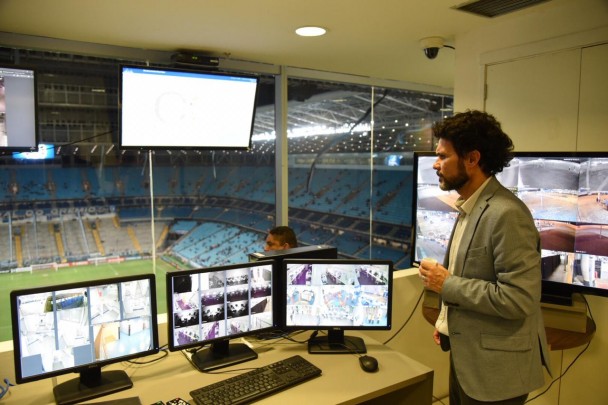Delegado Leonel Carivali observa monitores de vigilância na Arena do Grêmio. Ao fundo, o interior das arquibancada do estádio aparece pela janela da sala no piso superior do estádio.