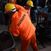 Homem com uniforme laranja no qual se lê às costas a palavra "Susepe" despeja massa de concreto em máquina para produção de blocos de construção.