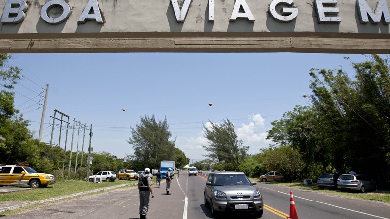 P&oacute;rtico de sa&iacute;da de cidade com a inscri&ccedil;&atilde;o "Boa Viagem" no topo da imagem. Abaixo estrada com carro sendo fiscalizado por policial militar.