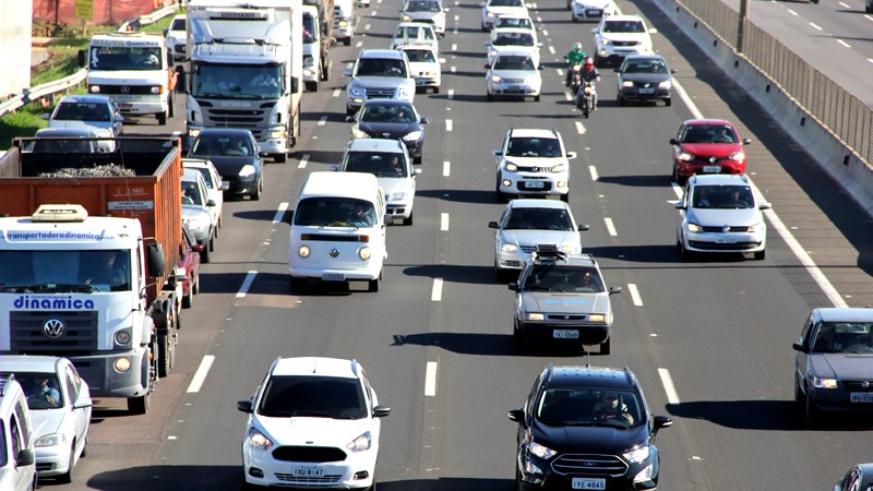 Imagem de uma pista de rodovia, com cinco faixas de rolagem no mesmo sentido, repleta de veículos trafegando durante o dia