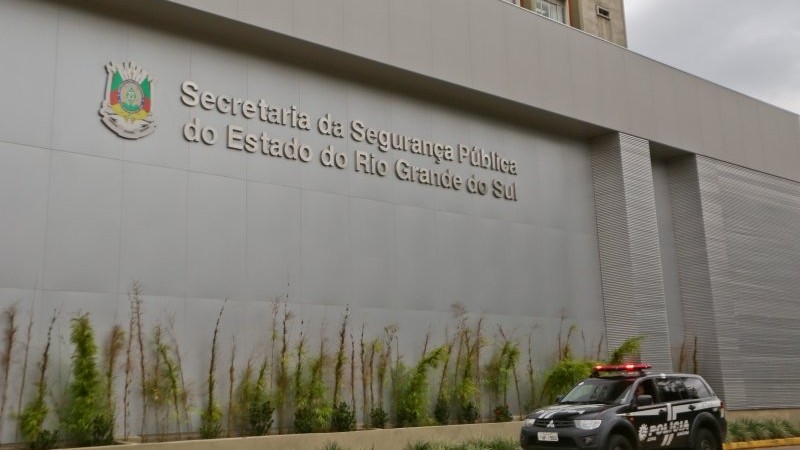 Nome da Secretaria da Segurança Pública do RS na fachada do prédio. Viatura da Polícia passando em frente ao prédio.