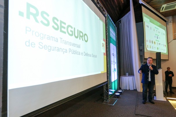 Ranolfo apresenta RS Seguro em Passo Fundo. Em primeiro plano, crescendo em profundidade até o centro da imagem, telão com a marca do RS Seguro. Ao fundo, na direita, Ranolfo em pé no palco.