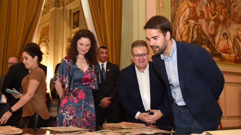 Diretora do IGP, Heloísa Küser, vice-governador, delegado Ranolfo Vieira Júnior, e governador Eduardo Leite observam documentos históricos sobre mesa no Salão Alberto Pasqualini, no Palácio Piratini.