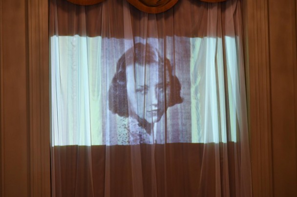Foto do rosto de Maria Luiza Häussler é projetada em cortina branca.