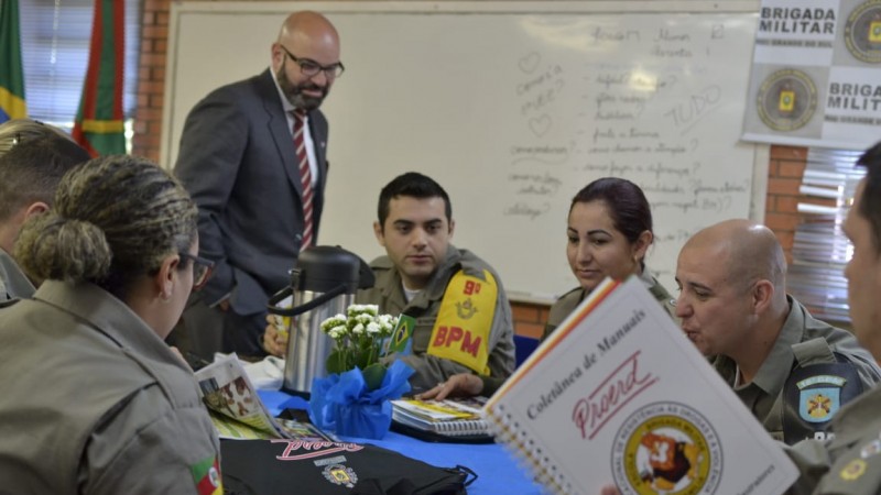 Aula do curso de formação de instrutores do PROERD. Policiais militares aparecem reunidos me torno de uma mesa. Um tome mate. Outro segura um caderno do Proerd. Ao fundo, lousa branca com anotações e banner da BM.