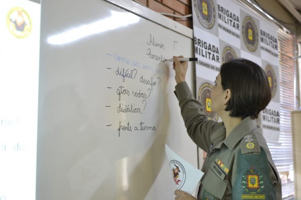 Policial militar feminina escreve com caneta piloto em uma lousa branca ao lado de um banner da Brigada Militar.