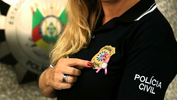Mulher com a camiseta da Polícia Civil aponta com o dedo indicador para a fita rosa que simboliza a campanha Outubro Rosa, presa à camiseta com botton da instituição, ao lado do brasão.