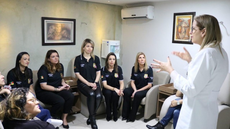 Grupo de policiais civis mulheres, entre as quais a chefe de Polícia, delegada Nadine Anflor, sentadas lado a lado em consultório onde uma médica vestindo jaleco branco fala sobre os cuidados preventivos contra o câncer de mama.