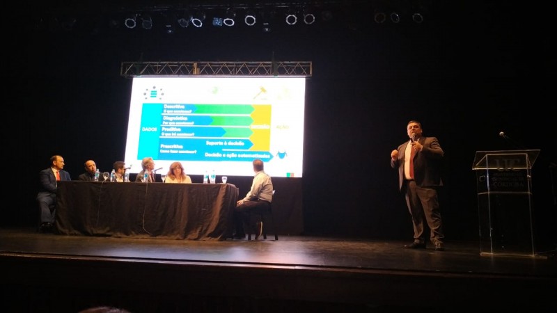 Major Donato aparece em palco de auditório lado de telão com apresentação. Também no palco, mesa com outros participantes de seminário.