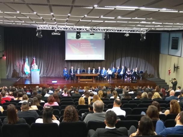 Plateia de auditório diante de palco com palestrantes de seminário sentados em cadeiras dispostas lado a lado do centro para a direita. À esquerda do palco, homem fala em púlpito. Telão mostra o nome do evento.