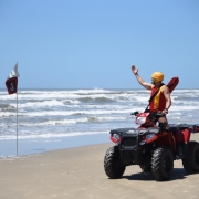 Na praia de Tramandaí, guarda-vida em cima de um quadriciclo faz sinal com o braço estendido em direção ao mar. Ao lado, uma bandeira de sinalização fincada na areia.