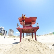 Visão frontal de uma guarita de praia, de onde um guarda-vidas salta em direção a um monte de areia no solo e aparece na imagem ainda no ar com os braços abertos. Ao fundo, céu azul e prédios da beira-mar.