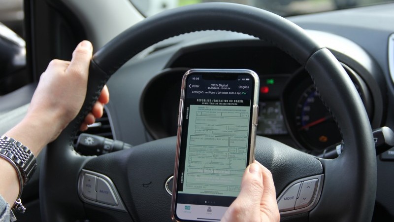 Mãos de um motorista dentro do carro seguram o volante e celular com o documento virtual