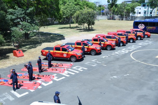 Fileira de bombeiros posicionados ao lado de viaturas estacionadas no pátio da Escola de Bombeiros, em Porto Alegre.