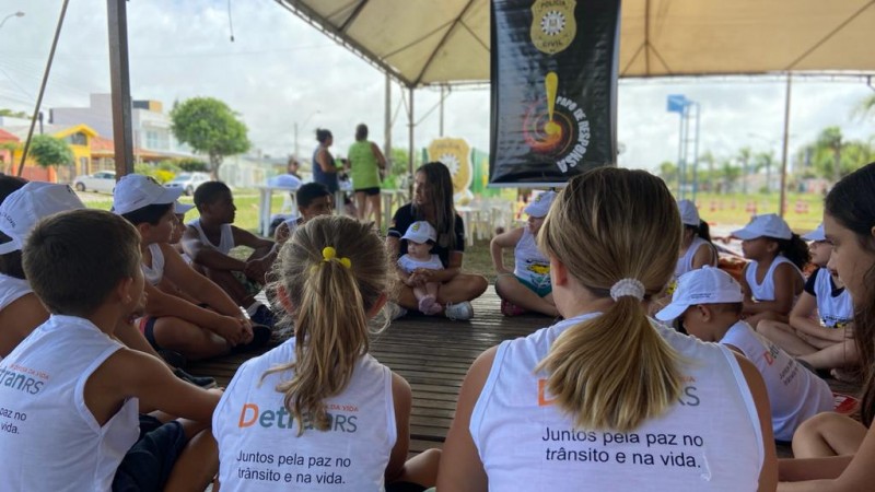 Crianças com camisetas patrocinadas pelo DetranRS sentadas em circulo acompanhados de uma policial civil. Ao fundo, banner do Papo de Responsa.