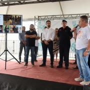 Vice-governador Ranolfo Vieira Júnior, de camisa branca da RS Verão Total, calça jeans e tênis, sobre um palco, observa telão com apresentados de dados da Operação. No palco, estão o governador Eduardo Leite e o comandante dos Bombeiros, cel. Bonfanti.