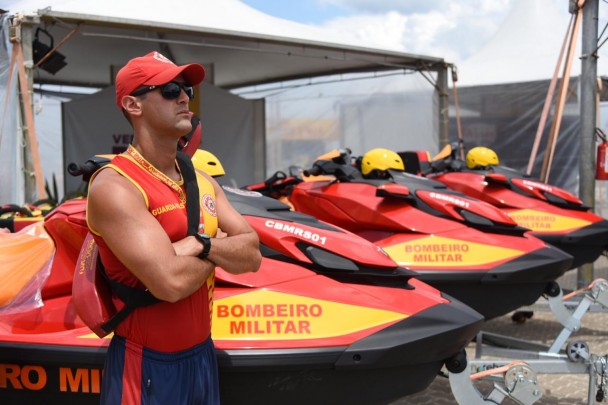 Guarda-vidas uniformizadas posa com os braços cruzados diante de três motos aquáticas com adesivagem do corpo de bombeiros, posicionadas uma ao lado da outra.