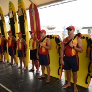 Nove guarda-vidas uniformizados posicionados um ao lado do outro. Cada um segura às costas uma prancha de salvamento amarela.