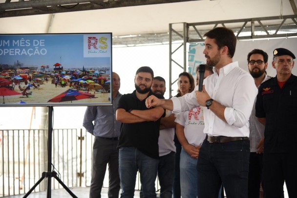 Governador Eduardo Leite, de camisa branca e calça jeans preta, fala ao microfone que segura com o braço esquerdo enquanto estende o braço direito. Ao lado, telão com a tela inicial de apresentação do balanço de um mês da Operação Verão.