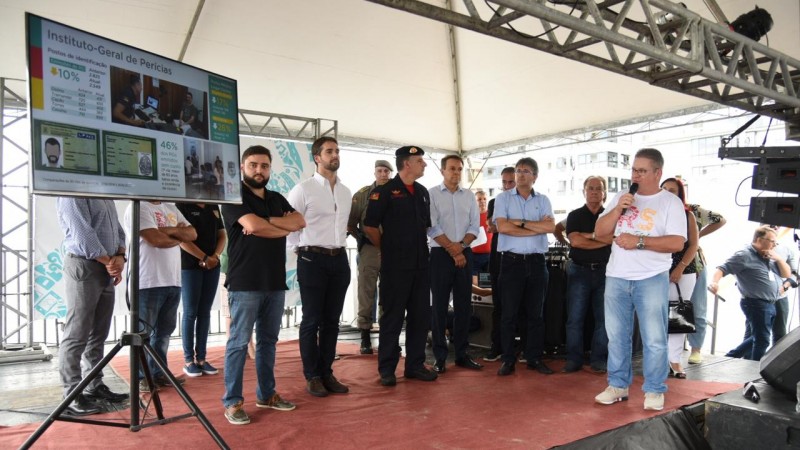 Em um palco montado na praia de Capão da Canoa, um telão com dados da Operação RS Verão Total no canto esquerdo da imagem. Ao centro, o governador e o comandante dos bombeiros. À direita, o vice-governador, que fala ao microfone.