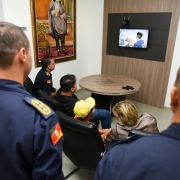 Em primeiro plano, dois bombeiros de costas. Ao fundo, família sentada ao lado do comandante do CBMRS observa TV com vídeo de homenagem aos guarda-vidas.