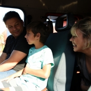 No interior da cabine de um caminhão do CBMRS, pai, filho e mãe, que observa o menino e sorri.