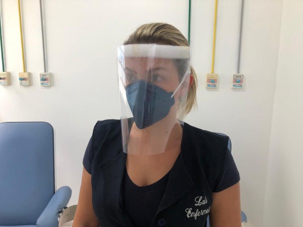 Profissional de saúde, mulher, aparece vestindo um protetor facial de acrílico transparente em frente ao rosto, preso na cabeça por uma tira de plástico branco.
