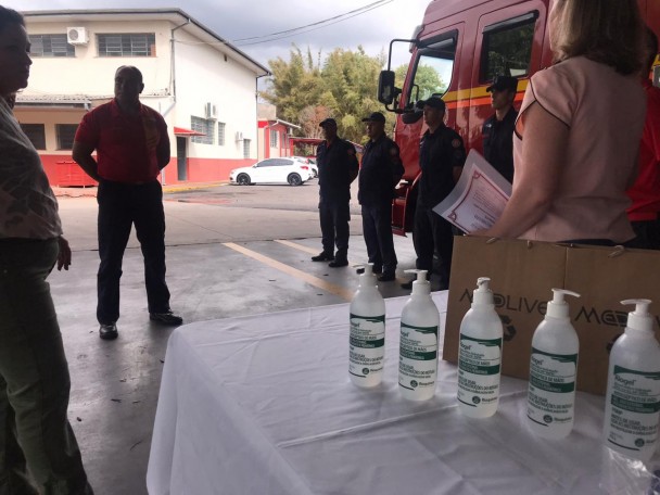 Frascos de álcool gel sobre uma mesa com toalha branca. Ao fundo, bombeiros conversam com representantes da empresa Medlive Distribuidora de Medicamentos.
