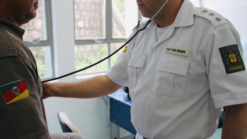 Oficial de saúde da BM, com farda branca, utiliza estetoscópio para auscultar o peito de outro brigadiano, de farda verde. Ambos estão em um consultório. O corte da imagem mostra os dois apenas da cintura pra cima e sem mostrar seus rostos.