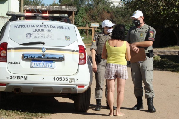 Ao lado de uma viatura da Patrulha Maria da Penha, dois policiais militares (uma mulher e um homem) conversam com uma mulher que aparece de costas em uma rua de chão batido.