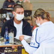 Servidora do Departamento de Saúde da BM confere documentação entregue por candidato a soldado da Brigada Militar. Os dois estão sentados, um de lado lado de uma mesa. Ambos usam máscaras.