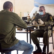 Policial militar entrevista candidato a aluno soldado da BM. Ambos usam máscaras e estão sendo frente a frente, cada um diante de uma mesa de classe escolar, dentro de um ginásio.