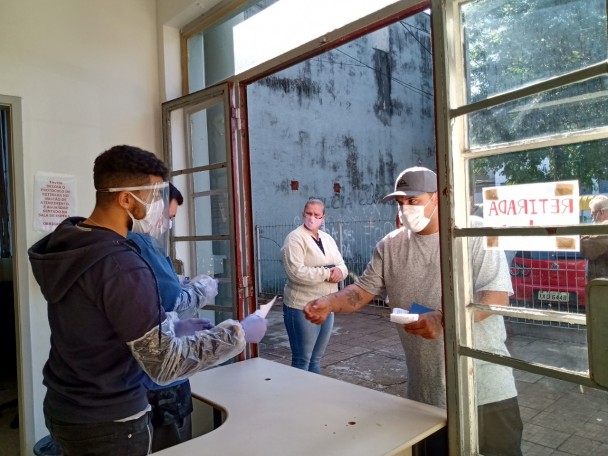 Servidor do IGP, usando equipamentos de proteção contra o coronavírus segura papel diante de um balcão com uma grande janela. Do outro lado, cidadão de máscara no rosto estende a mão para pegar o papel.