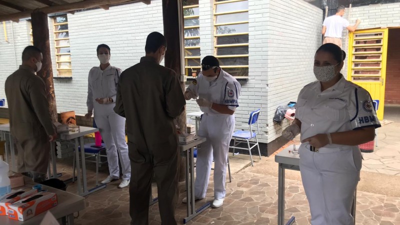 Sobre a cobertura de uma garagem, três profissionais do Departamento de Saúde da Brigada Militar, de farda branca, recebem dois alunos soldados, de macacão ocre, para realizar o teste da Covid-19.
