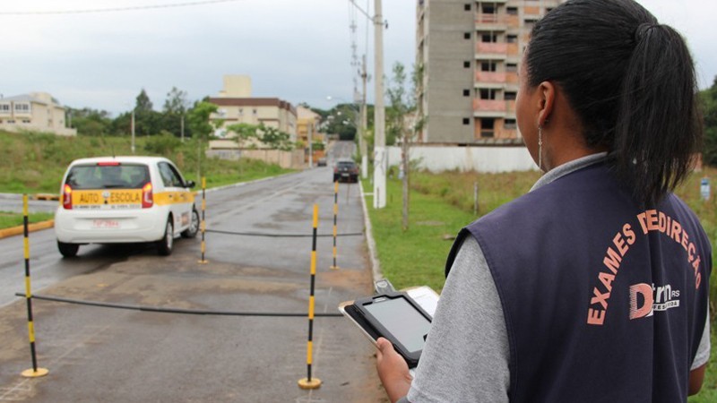 Examinadora do DetranRS, com uma tablet na mão, observa veículo de autoescola que se posiciona para fazer a baliza de estacionamento entre mastros listrados de amarelo e preto que demarcam a vaga em uma rua.