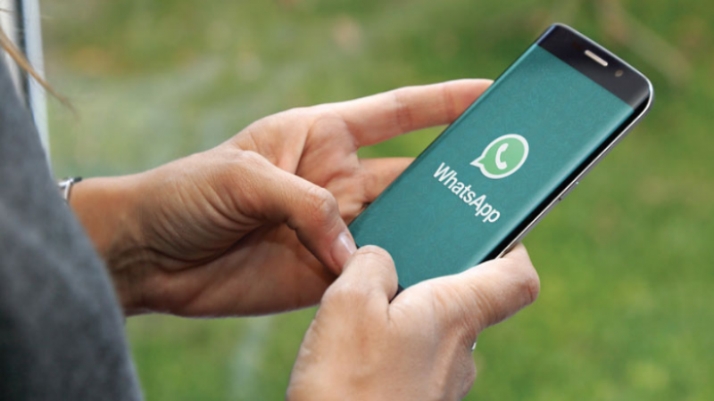 Imagem fechada em duas mãos de uma pessoa branca que segura um celular, em cuja tela aparece o logo do aplicativo WhatsApp.