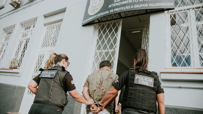 Duas policiais civis conduzem um homem algemado em direção à porta da DEAM de Porto Alegre.