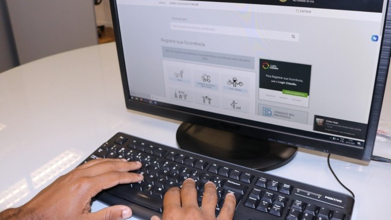 Mãos sobre um teclado de computador em cima de uma mesa, diante de um monitor, no qual aparece a capa do site da Delegacia Online da Polícia Civil.