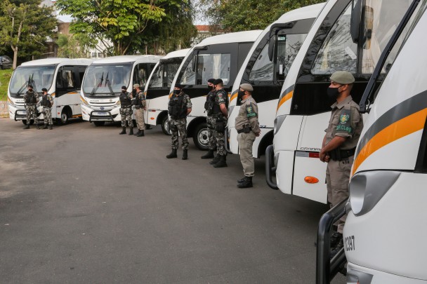 Sete micro-ônibus estacionados um ao lado do outro. Na frente de cada veículo, policiais militares em posição de descansar.