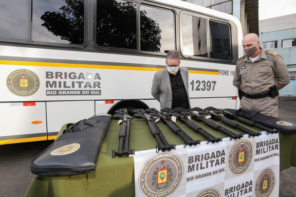 Vice-governador Ranolfo e comandante-geral da BM observam armas sobre uma mesa com banner da BM. Ao fundo, micro-ônibus da Brigada.