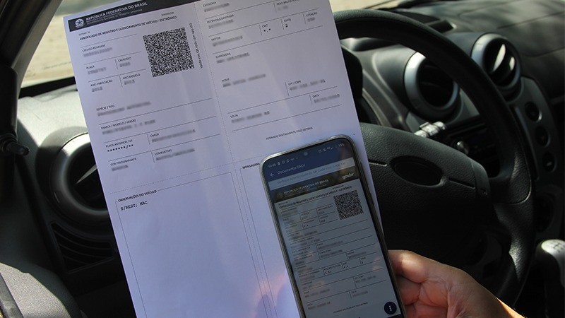 Foto mostra um pessoa sentada em frente ao volante de um carro segurando uma impressão do CRLV e um celular em cuja tela aparece a reprodução do documento.