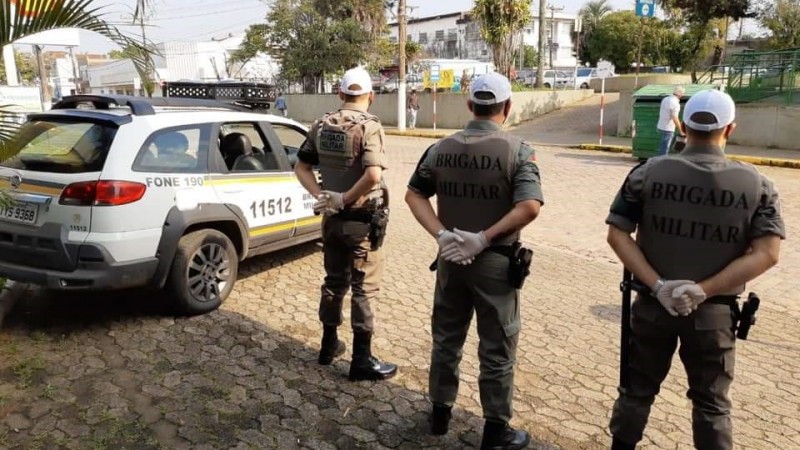 Três policiais militares aparecem de costas, em pé, em posição descansar, lado a lado e ao lado de uma viatura da Brigada Militar, diante de uma rua da cidade de Rio Pardo.